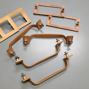 May include: A set of six gold-toned cabinet hardware pieces, including three rectangular pulls and three curved handles. The pulls have a simple, rectangular design with rounded corners, while the handles have a more elaborate, curved shape. All of the pieces have a brushed gold finish and feature small, silver-toned screws.