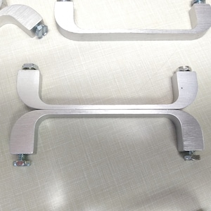 May include: Two silver metal handles with rounded edges and screw attachments. The handles are stacked on top of each other.