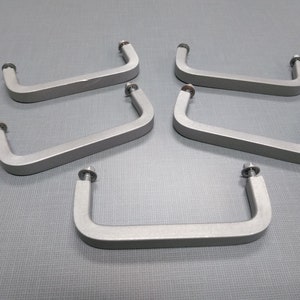 May include: Five silver-colored, rectangular cabinet pulls with rounded corners and attached bolts. The pulls are arranged on a gray surface, showcasing their design and hardware for furniture or cabinetry.