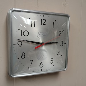 May include: A square wall clock with a silver metallic face and a silver frame. The clock features black Arabic numerals, black hour and minute hands, and a red second hand. The word Simplex is printed in black near the top center of the face.