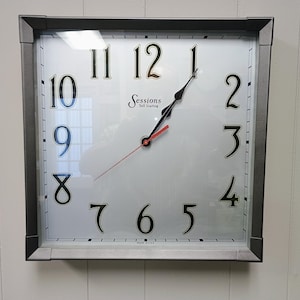 May include: Square wall clock with a silver frame and a white face. Black numerals mark the hours, with black and red hands indicating the time. The clock face has the text "Sessions" and "Self Starting".