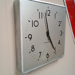 May include: A square wall clock with a brushed silver metal frame and a silver face. The clock features black Arabic numerals from 1 to 12, black hour and minute hands, and the text STROMBERG printed in black below the center. MADE IN USA is visible.