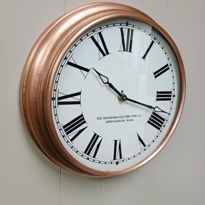 May include: A round wall clock with a copper-colored frame. The white face features black Roman numerals and hands. The text "THE STANDARD ELECTRIC TIME CO. SPRINGFIELD, MASS." is printed on the face.