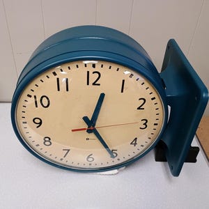 May include: A double-sided, teal-colored clock with a cream-colored face. The clock has black numerals and hands, with a red second hand. The clock is mounted on a teal bracket, and is likely for a business or public space.