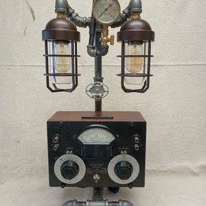 May include: A steampunk-style lamp with two caged light bulbs, a gauge, and a vintage-looking radio panel with dials and knobs. The lamp is made of metal pipes and has a wooden base.
