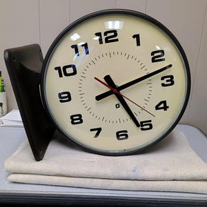 May include: A double-sided wall-mounted clock with a dark brown frame and a cream-colored face. The clock features bold black Arabic numerals from 1 to 12, black hour and minute hands, and a thin red second hand. A small logo is visible at the center.
