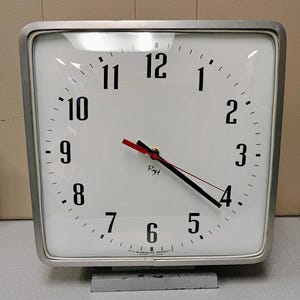 May include: A white square clock with black hands and black numbers. The clock face has a red second hand and the text "Ryt" in the center. The clock is mounted on a silver stand.