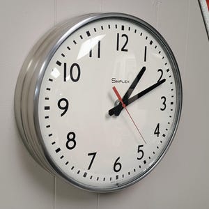 May include: A round, silver-framed wall clock with a white face. Black numerals mark the hours, with a red second hand and black hour and minute hands. The clock face reads "SIMPLEX".
