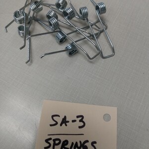 May include: A close-up of a pile of silver metal springs. A white note with black writing reads "SA-3 SPRINGS".