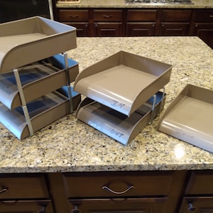 May include: Four beige plastic desk organizers with metal frames. The organizers are stacked in two sets of two. The top organizer of each set has handwritten labels: "In" and "Out". The bottom organizer of the set of two has a handwritten label: "To File".