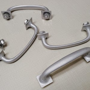 May include: Five silver metal cabinet door handles with square mounting plates and screws. The handles are all in the shape of a loop.