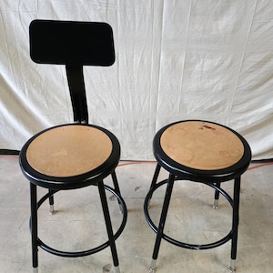 May include: Two black metal stools with round, light brown seats. One stool has a black backrest. The stools have a circular footrest and silver-colored feet. The stools are on a concrete floor.