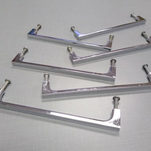May include: Six chrome-finished cabinet pulls with mounting screws. Each pull has a rectangular bar with a curved end and a screw post. The pulls are arranged on a light gray surface.
