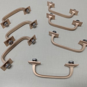 May include: Ten gold-toned cabinet pulls with a simple, curved design. Each pull has a square base with a single screw hole.