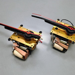 May include: Two clock mechanisms featuring square gold-toned metal plates, black textured hands, and thin red pointers. Each unit includes a copper-colored coil component with a white cylindrical core and black mounting screws at the corners.