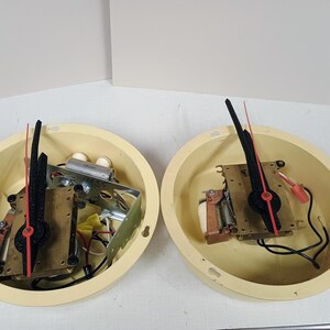 May include: Two circular cream-colored clock housings containing exposed internal mechanisms. Each unit features a central brass-toned plate, a black hour and minute hand, and a thin red second hand. Electrical wiring and small components are visible.
