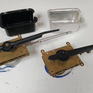 May include: Two brass clock mechanisms with long black textured hands and blue electrical wires, accompanied by one black rectangular plastic housing and one silver-colored metallic rectangular housing, all displayed as separate components.