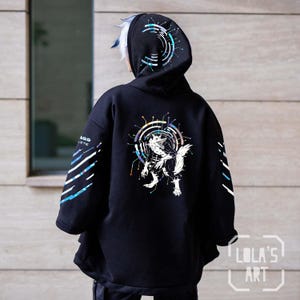 Może przedstawiać: Czarna bluza z kapturem z holograficznym wzorem wilka w kółku z obwodami na plecach. Rękawy mają biało-niebieskie paski. Tekst "LOLA'S ART" znajduje się w prawym dolnym rogu bluzy.