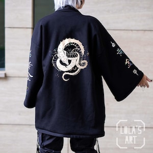 Könnte beinhalten: Schwarze Jacke im Kimono-Stil mit einem großen, verzierten weißen und goldenen Drachendesign auf dem Rücken. Die Ärmel zeigen farbenfrohe japanische Schriftzeichen. Die Jacke hat eine lockere Passform und einen kragenlosen Schnitt. "Lola's Art" ist unten rechts aufgedruckt.