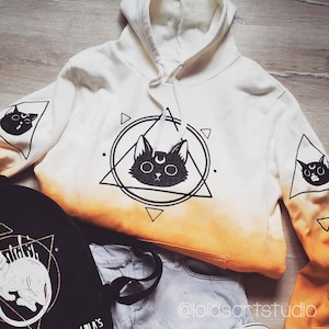 Puede incluir: Sudadera blanca con efecto degradado naranja. La sudadera presenta un gato negro con una luna creciente en la cabeza dentro de un círculo y un diseño triangular. El texto "@lolasartstudio" es visible en la esquina inferior derecha.