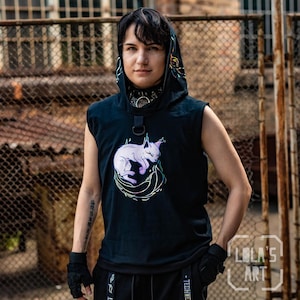 Puede incluir: Una camiseta negra sin mangas con capucha que presenta un gráfico de un gato de color morado claro acurrucado en un nido verde brillante. La capucha tiene un diseño de gato a juego y el cuello incluye un detalle de anillo en D negro.