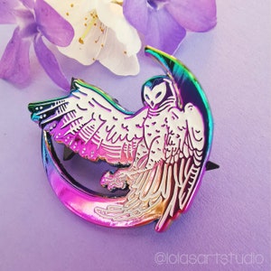 May include: A colorful enamel pin featuring an owl perched on a crescent moon. The pin has a metallic rainbow finish with white and iridescent details. The owl's wings are spread, and the design is set against a purple background with flowers.