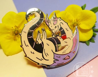 May include: A gold-toned metal enamel pin featuring a cream-colored dragon curled into a crescent moon shape. The dragon has pink accents on its ears and tail, with a purple crescent base. The pin includes a small logo reading Lola's Art.