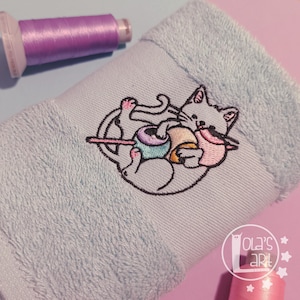 Op de afbeelding: Lichtblauwe handdoek met een geborduurd kattenontwerp met kleurrijke bollen garen. Een spoel lila garen staat linksboven en een roze spoel rechtsonder. De handdoek heeft een zachte, getextureerde afwerking. Ontwerp van Ola's Art.