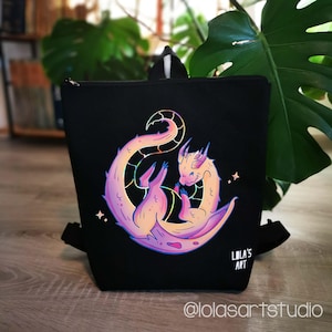 Könnte beinhalten: Ein schwarzer Rucksack mit einer Grafik eines zusammengerollten Drachen in Rosa-, Orange- und Gelbtönen. Der Drache hat einen langen Schwanz und kleine Flügel. Der Text LOLA'S ART ist in Weiß unten rechts auf der Vorderseite aufgedruckt.