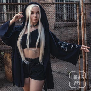 Könnte beinhalten: Schwarze Kapuzen-Kimono-Jacke mit weiten Ärmeln, verziert mit einem weißen Grafikdesign auf dem Ärmel. Das Model trägt ein schwarzes Crop-Top und Shorts. Der Hintergrund zeigt einen Maschendrahtzaun.