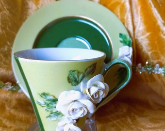 May include: A delicate teacup and saucer set with a green and white floral design. The teacup features a raised 3D floral design with white roses and green leaves.