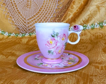 May include: A delicate pink teacup and saucer set with a floral design. The cup features a hand-painted design of pink roses with green leaves. The saucer has a gold rim and a matching floral design.