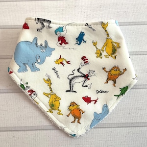 May include: White, triangular baby bib featuring a colorful Dr. Seuss print. The bib showcases various characters from the beloved children's books, including the Cat in the Hat and the Grinch. The bib has a soft, white trim.
