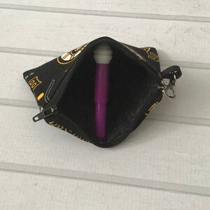 May include: A small black zippered pouch with a gold and white print of musical instruments. The pouch is open and a purple pen is inside.