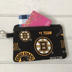 May include: Black zippered pouch with a Boston Bruins team print. The pouch has a gold zipper and a gold key chain. The pouch is filled with two gift cards.