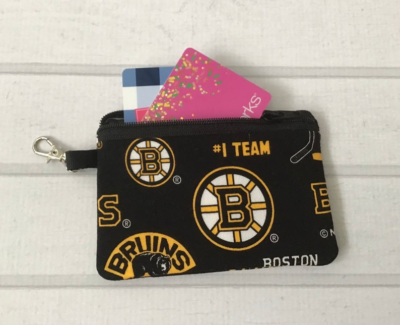 May include: Black zippered pouch with a Boston Bruins team print. The pouch has a gold zipper and a gold key chain. The pouch is filled with two gift cards.