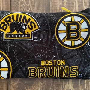 May include: A black and yellow Boston Bruins zipped pouch. The pouch displays the Bruins logo and team name in yellow and white on a black background. A small yellow loop and silver clasp are attached.