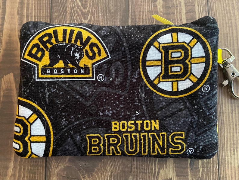 May include: A black and yellow Boston Bruins zipped pouch. The pouch displays the Bruins logo and team name in yellow and white on a black background. A small yellow loop and silver clasp are attached.