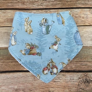 May include: A light blue fabric bib with a Peter Rabbit design. The bib features multiple images of Peter Rabbit and other characters from the book, including a watering can, carrots, and a blue bunny.
