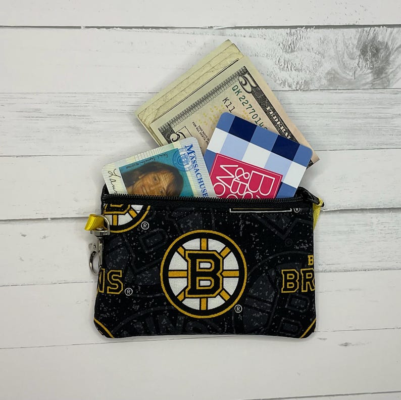 May include: Black coin purse with a Boston Bruins logo, containing cash and cards. The purse has a repeating pattern of the Bruins logo and text. The zipper pull is yellow.