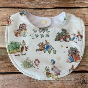 May include: A white baby bib with a charming Peter Rabbit print. The bib features a snap closure and is decorated with illustrations of rabbits, owls, and other woodland creatures in a garden setting. The bib is made of soft fabric.