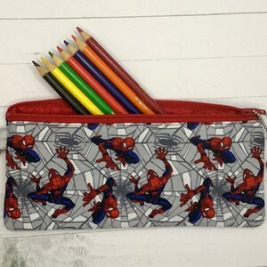 May include: A rectangular fabric pencil case featuring a repeating pattern of the character Spider-Man against a gray spiderweb background. The case has a red zipper closure, a silver metal lobster clasp on the side, and a hexagonal charm.