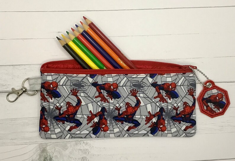 May include: A rectangular fabric pencil case featuring a repeating pattern of the character Spider-Man against a gray spiderweb background. The case has a red zipper closure, a silver metal lobster clasp on the side, and a hexagonal charm.