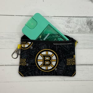 May include: A small black zippered pouch with a Boston Bruins logo pattern, holding a teal spray bottle. The pouch has a yellow zip pull and a silver clasp. The Bruins logo is black, gold, and white.