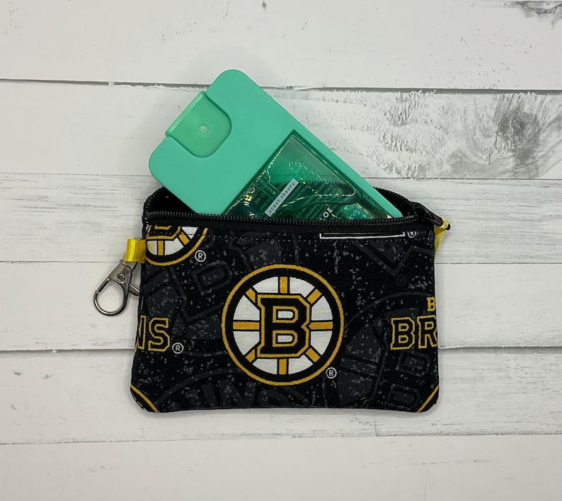 May include: A small black zippered pouch with a Boston Bruins logo pattern, holding a teal spray bottle. The pouch has a yellow zip pull and a silver clasp. The Bruins logo is black, gold, and white.
