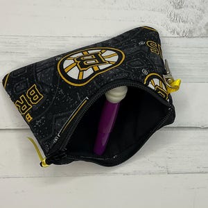 May include: Black zippered pouch with the Boston Bruins logo in yellow and white on a black background. The pouch is open, showing a purple object. The zipper pull is yellow.