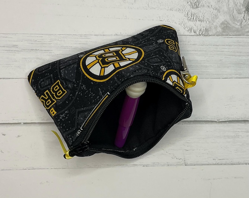 May include: Black zippered pouch with the Boston Bruins logo in yellow and white on a black background. The pouch is open, showing a purple object. The zipper pull is yellow.