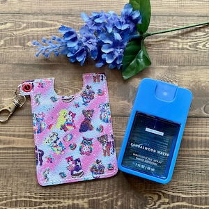 May include: A pink and blue key chain with a small, clear plastic bottle attached. The bottle is labeled "Sandalwood Waves Moisturizing Spray Hand Sanitizer 1 fl oz / 30 ml".