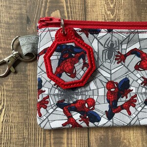 May include: A rectangular fabric pouch featuring a repeating pattern of the superhero Spider-Man in red and blue against a gray spiderweb background. The pouch has a red zipper and a matching red octagonal charm attached to a silver metal clip.