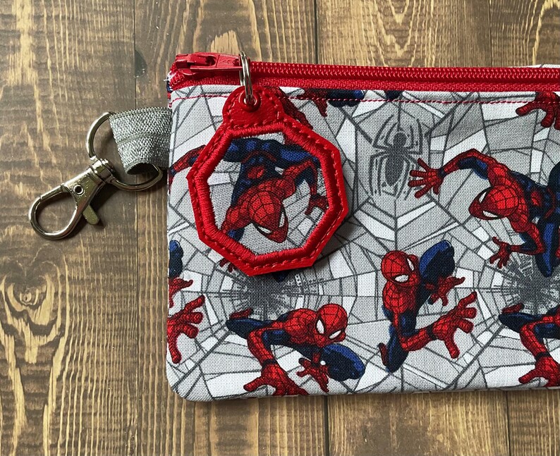 May include: A rectangular fabric pouch featuring a repeating pattern of the superhero Spider-Man in red and blue against a gray spiderweb background. The pouch has a red zipper and a matching red octagonal charm attached to a silver metal clip.